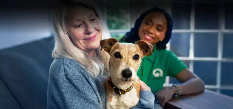 An older woman sits on a sofa holding her small rescue pet dog, while a smiling carer in a green shirt sits beside her. Natural light enters through a window in the background.