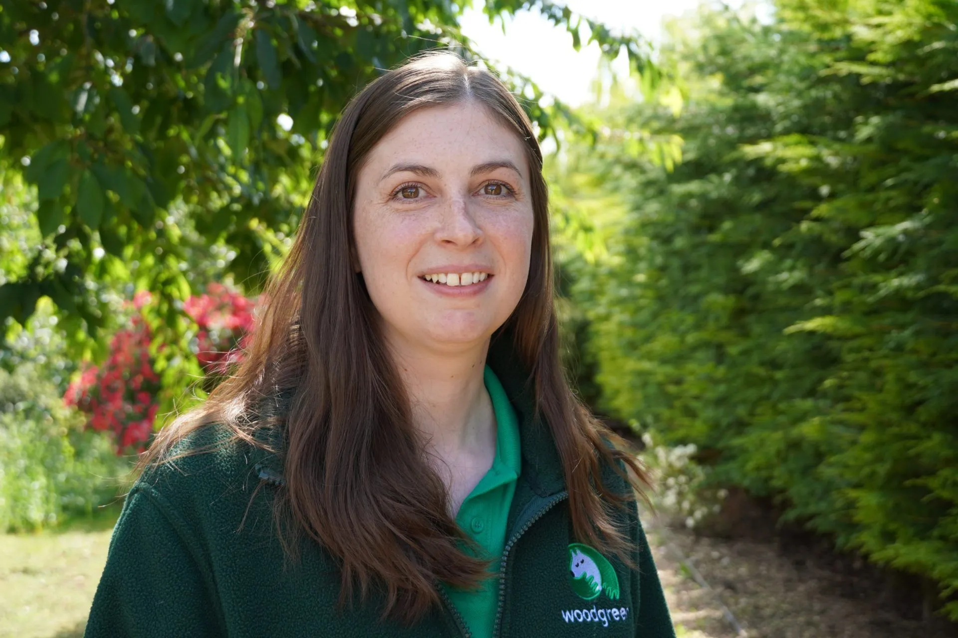 Sam Ryan, a woman with long brown hair, stands outdoors smiling. She is wearing a green fleece with a Woodgreen logo and a green polo shirt. Lush trees and greenery form the background on this sunny day.