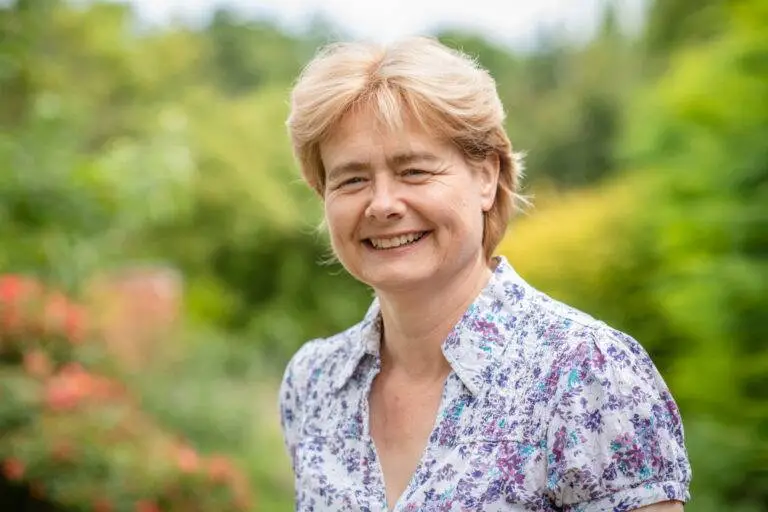 Wendy Kruger, with short light brown hair, smiles outdoors in a white shirt adorned with a blue and purple floral pattern. The lush green background features blurred trees and bushes.
