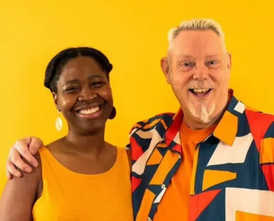 Two people smiling and standing close together against a bright yellow background. One wears a yellow dress and white earrings, and the other wears a multicolored shirt with orange and yellow accents.