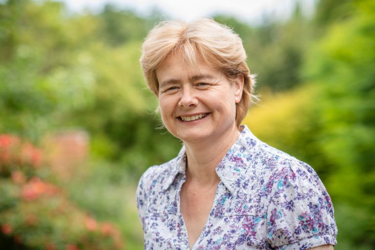 Wendy Kruger, with short light brown hair, smiles outdoors in a white shirt adorned with a blue and purple floral pattern. The lush green background features blurred trees and bushes.