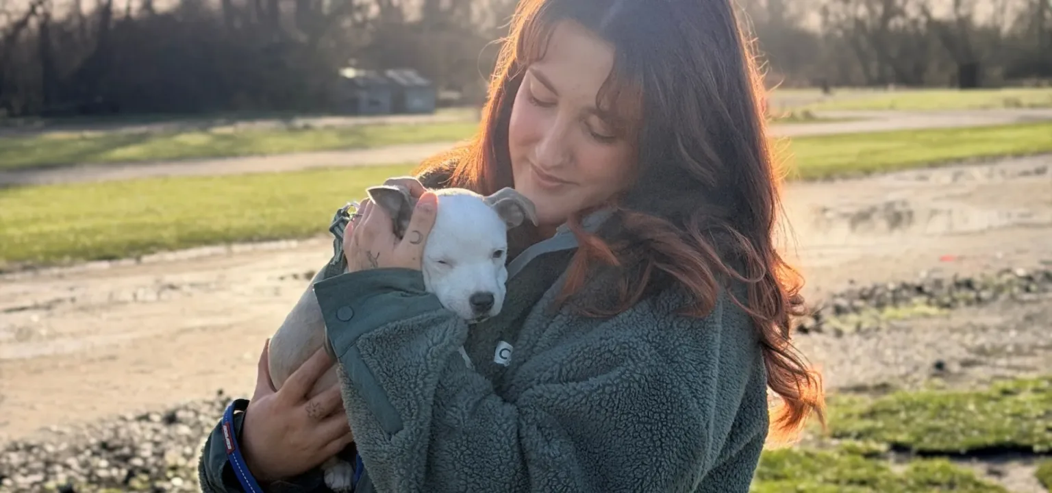 A woman with long brown hair holds a small light grey and white puppy outdoors on a sunny day, with green grass and trees in the background. Both appear calm and content.