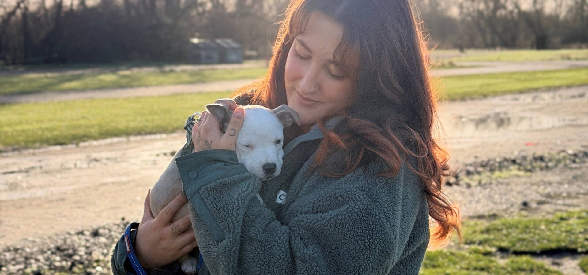 A woman with long brown hair holds a small light grey and white puppy outdoors on a sunny day, with green grass and trees in the background. Both appear calm and content.