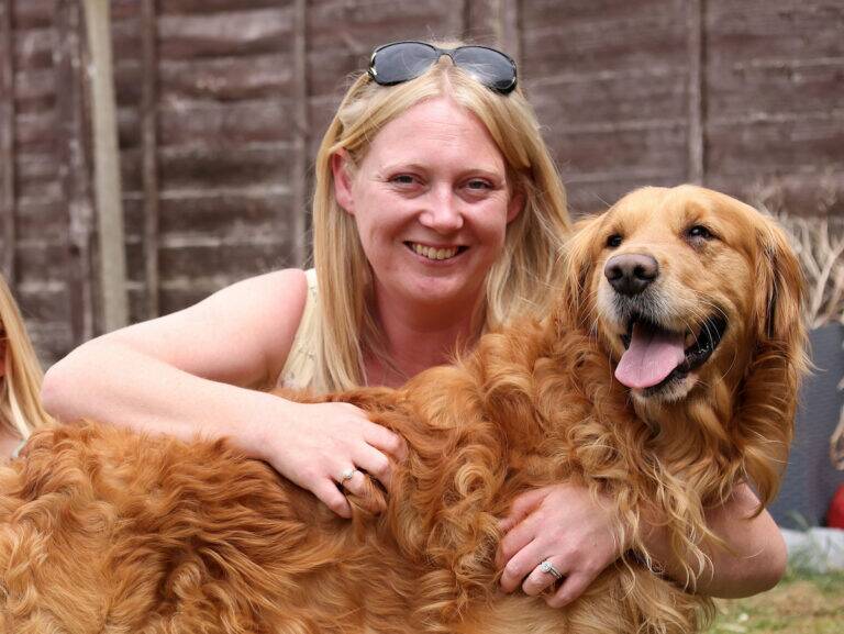 A woman with blonde hair and sunglasses on her head smiles while hugging a happy golden retriever outdoors, with a wooden fence in the background.