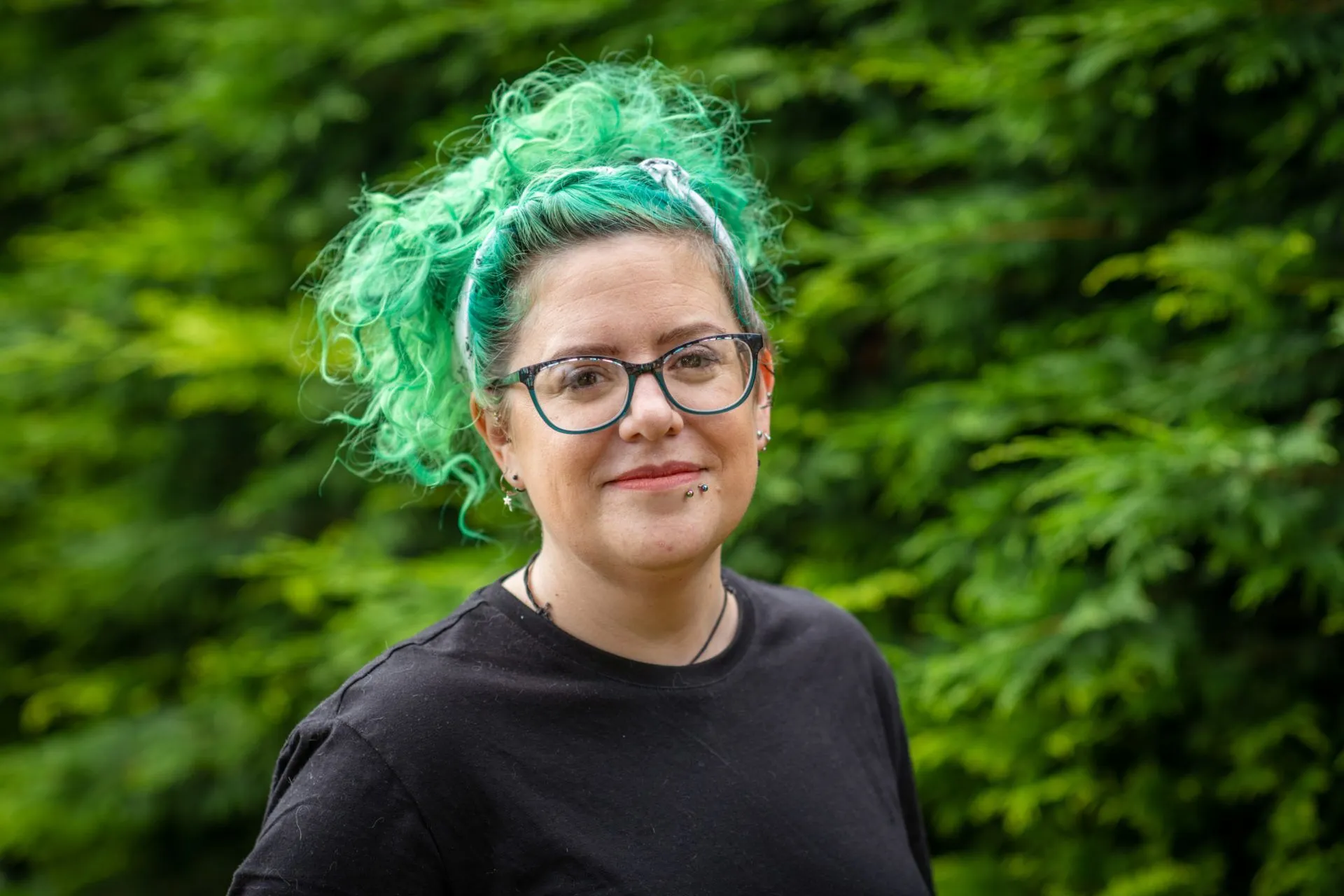 Sam Prior, a person with curly green hair, and glasses, stands outdoors in front of lush green foliage, wearing a black shirt and smiling slightly at the camera.