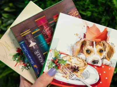 A hand with purple nail polish holds three festive holiday cards, one featuring a watercolor dog in a Santa hat with a mince pie, and another decorated with Christmas cracker illustrations—perfect finds from Woodgreen charity shops.
