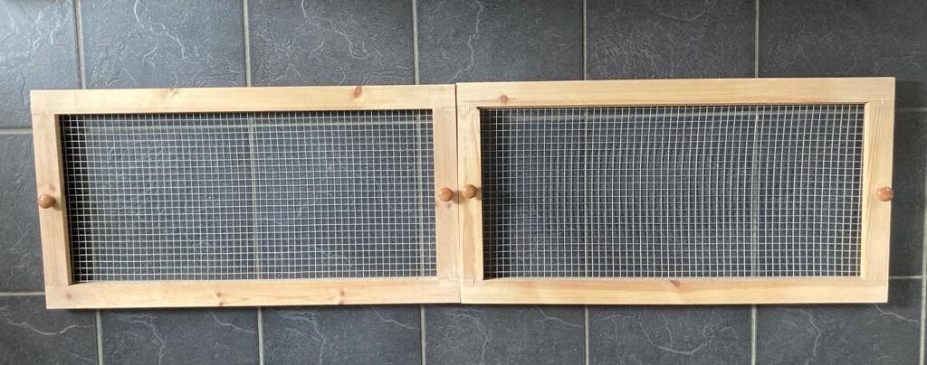 Two rectangular wooden frames with mesh wire centers, ideal as a gerbil homemade lid for tank, are joined side by side. Each features a small round wooden knob and is placed against a dark tiled wall.