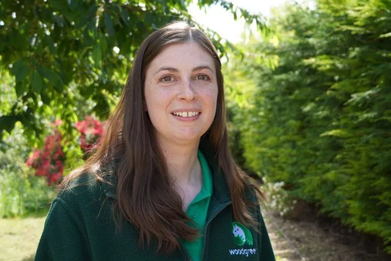 Sam Ryan, a woman with long brown hair, stands outdoors smiling. She is wearing a green fleece with a Woodgreen logo and a green polo shirt. Lush trees and greenery form the background on this sunny day.