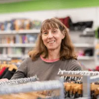 A woman with long brown hair and a gray shirt smiles while standing among clothing racks in a brightly lit store.