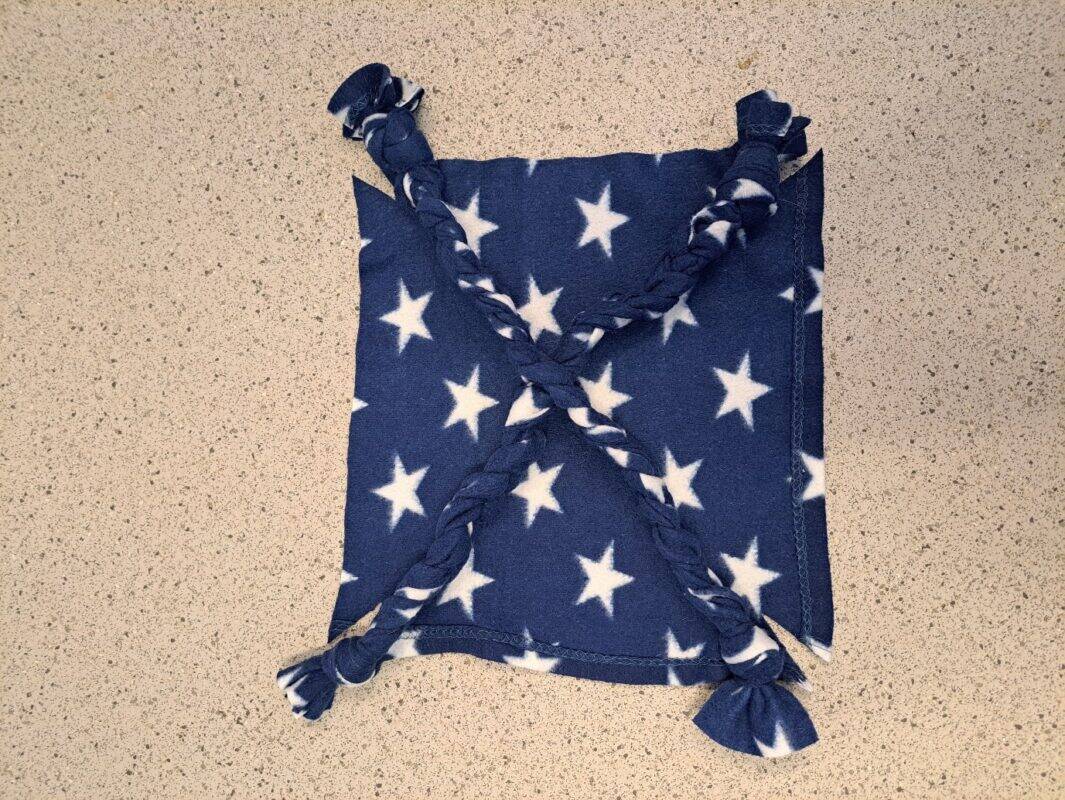 A square rat hammock made of navy blue fleece with white stars, featuring four braided corners tied together diagonally, placed on a speckled beige countertop.