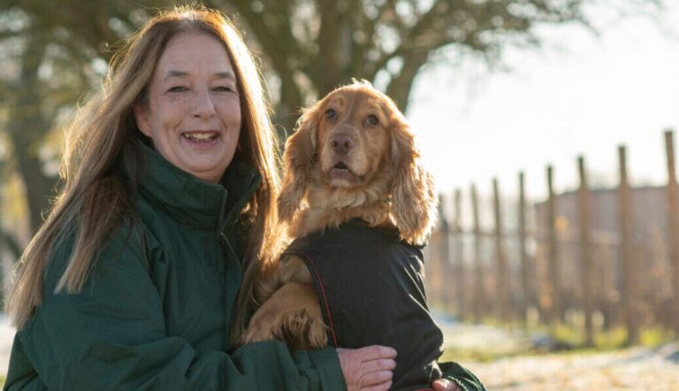 Sue Ketland, in a green jacket, smiles while holding a brown dog wearing a black coat outdoors on a sunny day, with trees and a fence in the background.