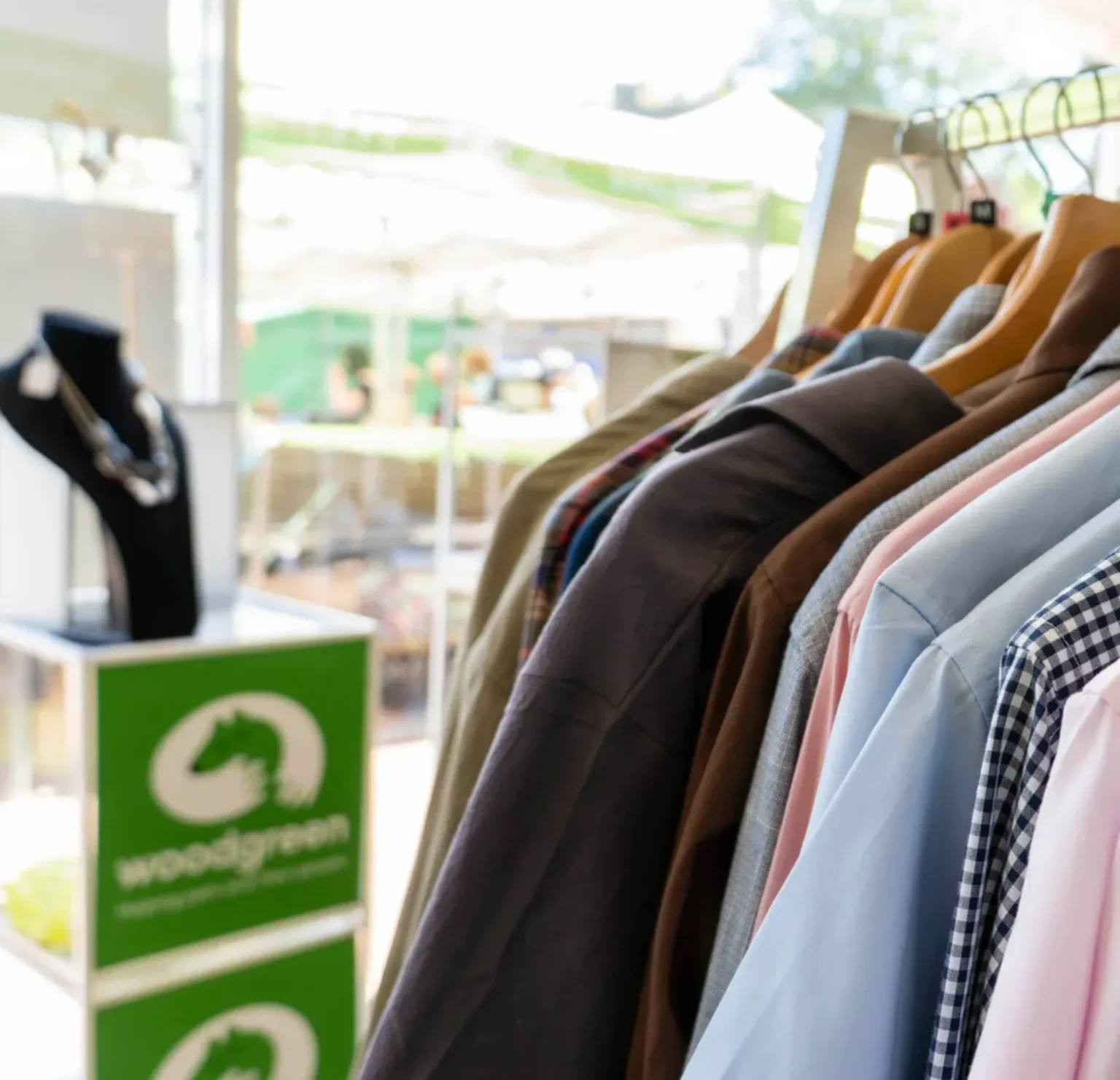 A rack of assorted shirts on hangers is displayed near a window in one of the Woodgreen charity shops, with a jewelry stand and green Woodgreen signs in the background. Sunlight shines through, creating a bright and inviting atmosphere.