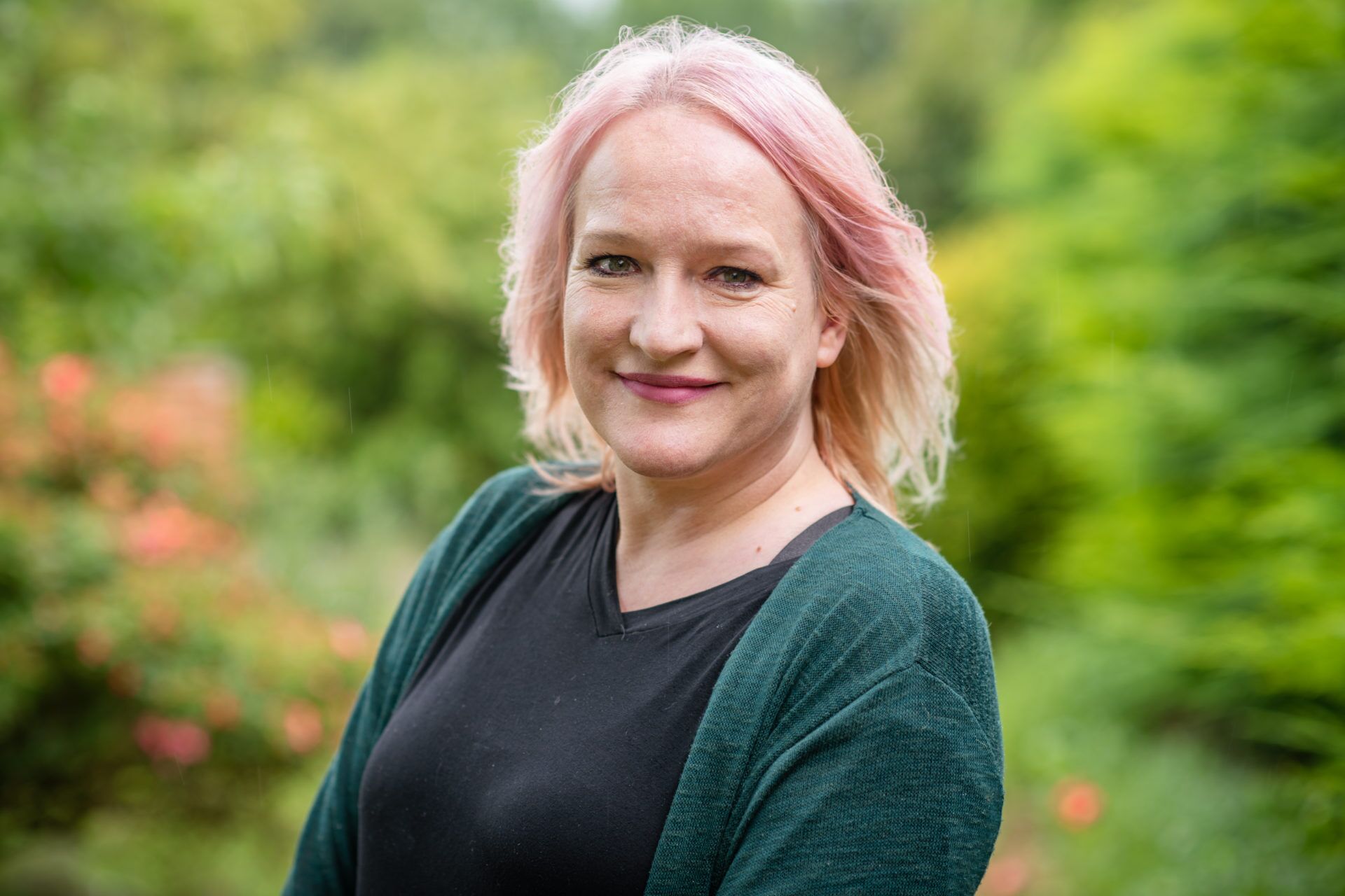 Juliette Jones, with light pink hair, smiles outdoors in a black shirt and dark green cardigan against a blurred background of green foliage and colorful flowers.