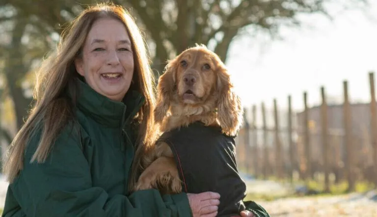 Sue Ketland, in a green jacket, smiles while holding a brown dog wearing a black coat outdoors on a sunny day, with trees and a fence in the background.