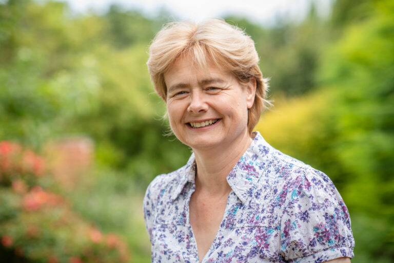 Wendy Kruger, with short light brown hair, smiles outdoors in a white shirt adorned with a blue and purple floral pattern. The lush green background features blurred trees and bushes.