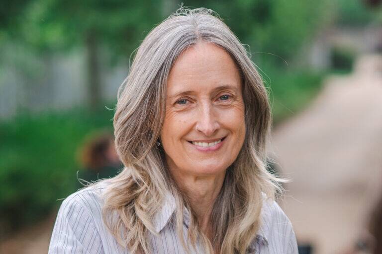 Wendy Jones, a woman with a striped shirt, smiles outdoors with greenery and a blurry path in the background.