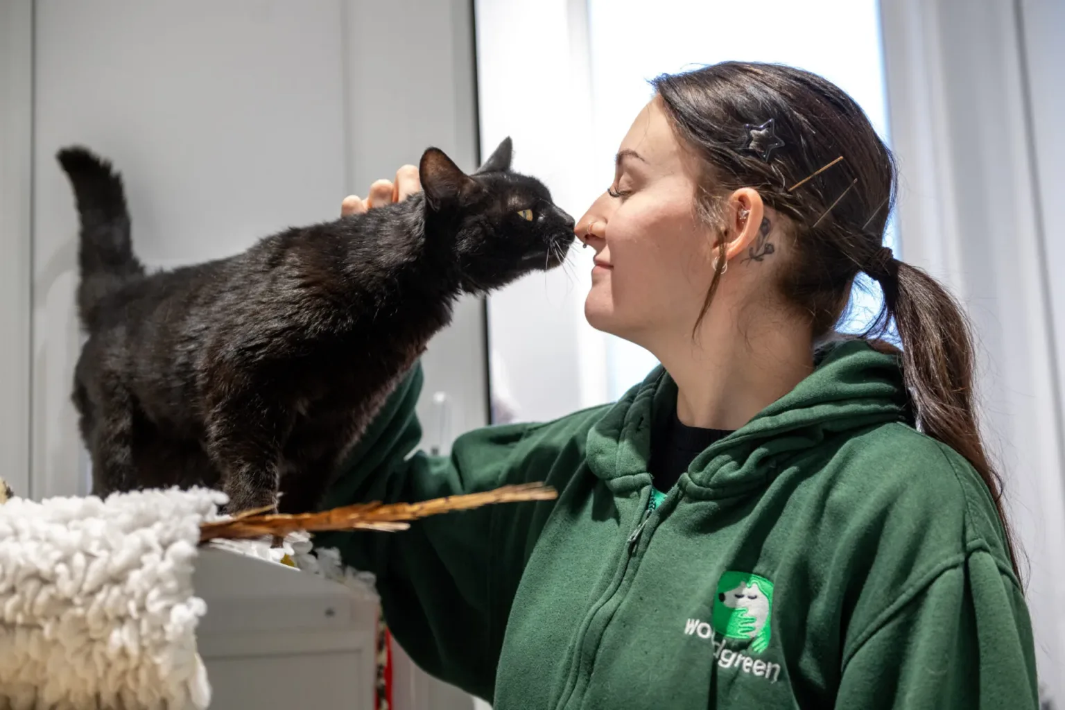 A Woodgreen carer in a green hoodie smiles as a black cat touches her nose with its own while she gently pets the cat. They appear to be enjoying a tender moment indoors.