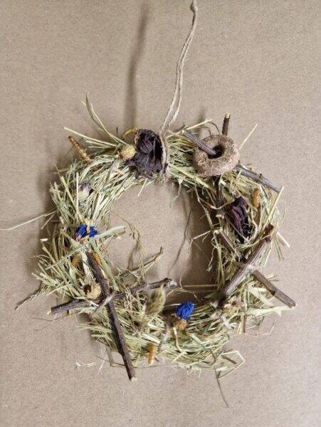A small, rustic wreath for rabbits and rodents, made of dried grass, twigs, and wildflowers, with a loop of string for hanging, displayed on a plain brown background.