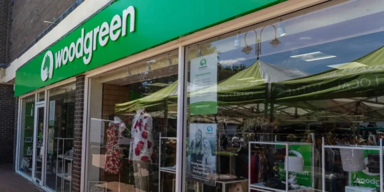 Storefront of a Woodgreen charity shop in Ely, featuring a green sign, clothing displays in the window, and outdoor market tents reflected in the glass.