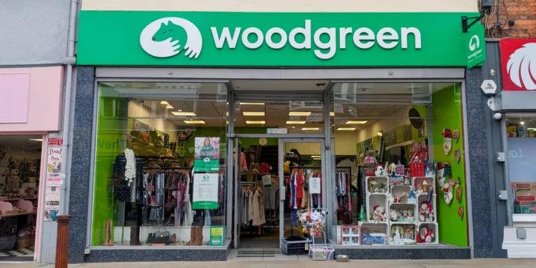 Storefront of a Woodgreen Kettering charity shop with a green sign, glass windows displaying clothes on racks, shelves with toys and ornaments, and a bright, inviting interior.