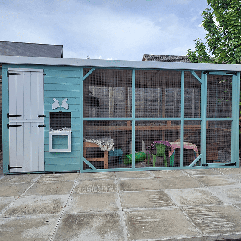 A large outdoor rabbit enclosure with a blue wooden structure, white door, mesh walls, and shelves inside. There are toys and blankets visible through the mesh. The enclosure sits on a paved patio.