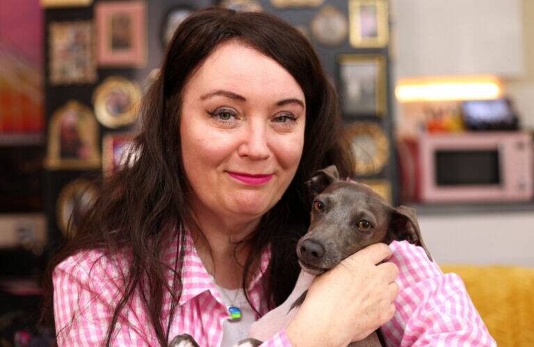 A woman with long dark hair in a pink checked shirt smiles while holding a small grey dog. The background shows framed photos and objects on a wall, and a pink microwave.