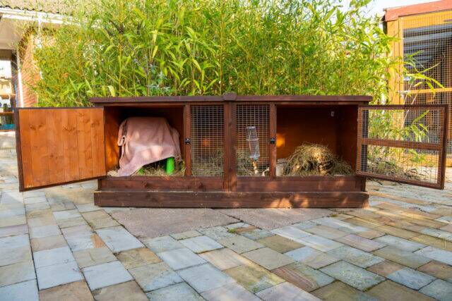A large wooden guinea pig hutch with open doors, revealing straw bedding and a blanket inside. The hutch sits on a stone patio, surrounded by green plants in the background.