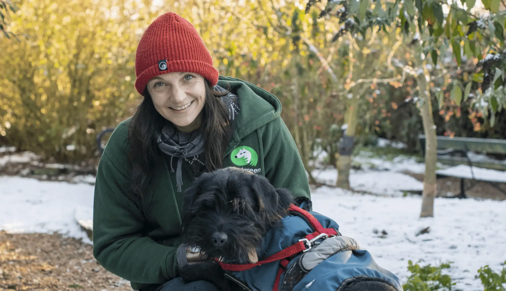 Britt Rosendahl, wearing a red knit hat and green jacket, smiles while kneeling next to a black dog in a blue coat. They are outdoors in a snowy, wooded area with trees and a bench in the background.