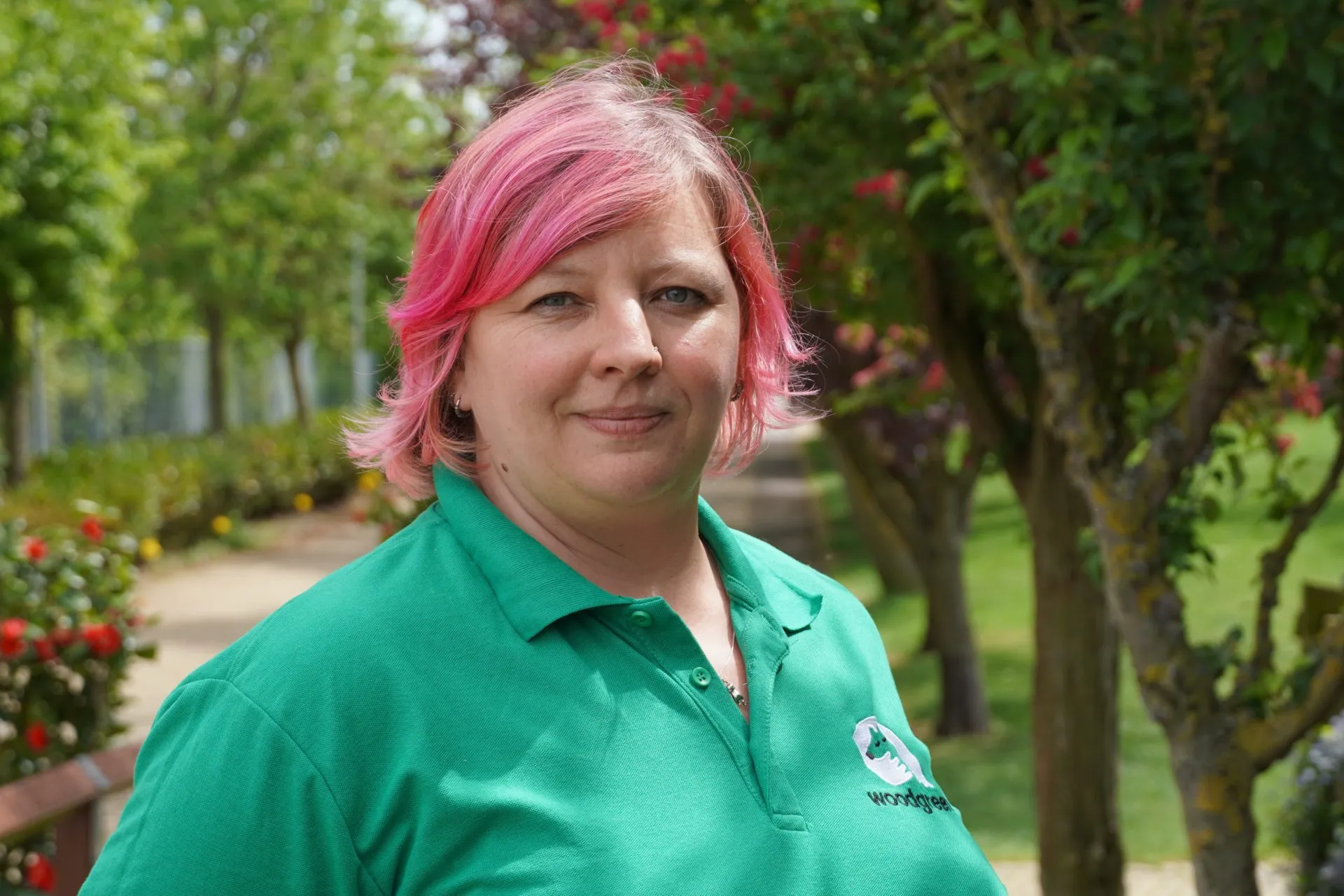 Lindsay Arliss with short pink hair and a green polo shirt stands outside in a garden with green trees and flowering bushes, smiling at the camera—a trusted pet expert for comment.