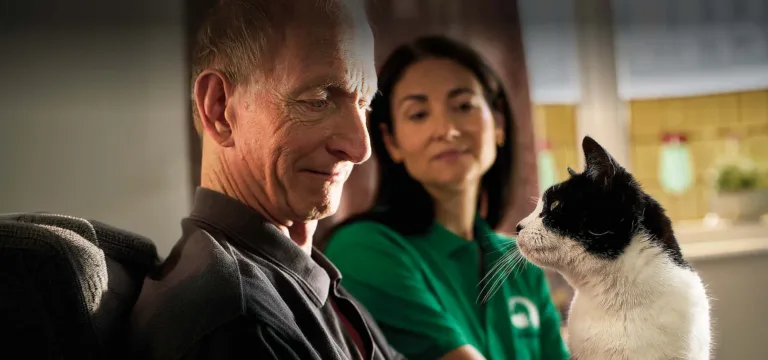 An elderly man smiles at a black and white cat on his lap, whilst a carer in a green shirt sits beside them, watching the heartwarming interaction—showcasing the joy that Woodgreen pets charity brings to people and their pets.
