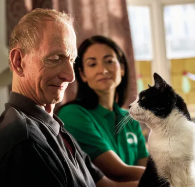 An older man smiles at a black and white cat sitting in front of him, while a woman in a green shirt, from Woodgreen pets charity, watches them, all indoors with soft, natural light.