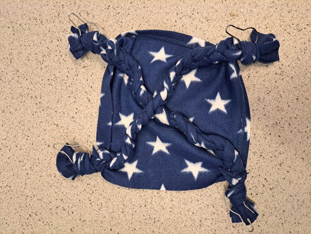 A square piece of blue fleece fabric with white stars, knotted and braided at the corners, forms a cozy rat hammock with metal hooks attached to each corner, lying on a speckled countertop.