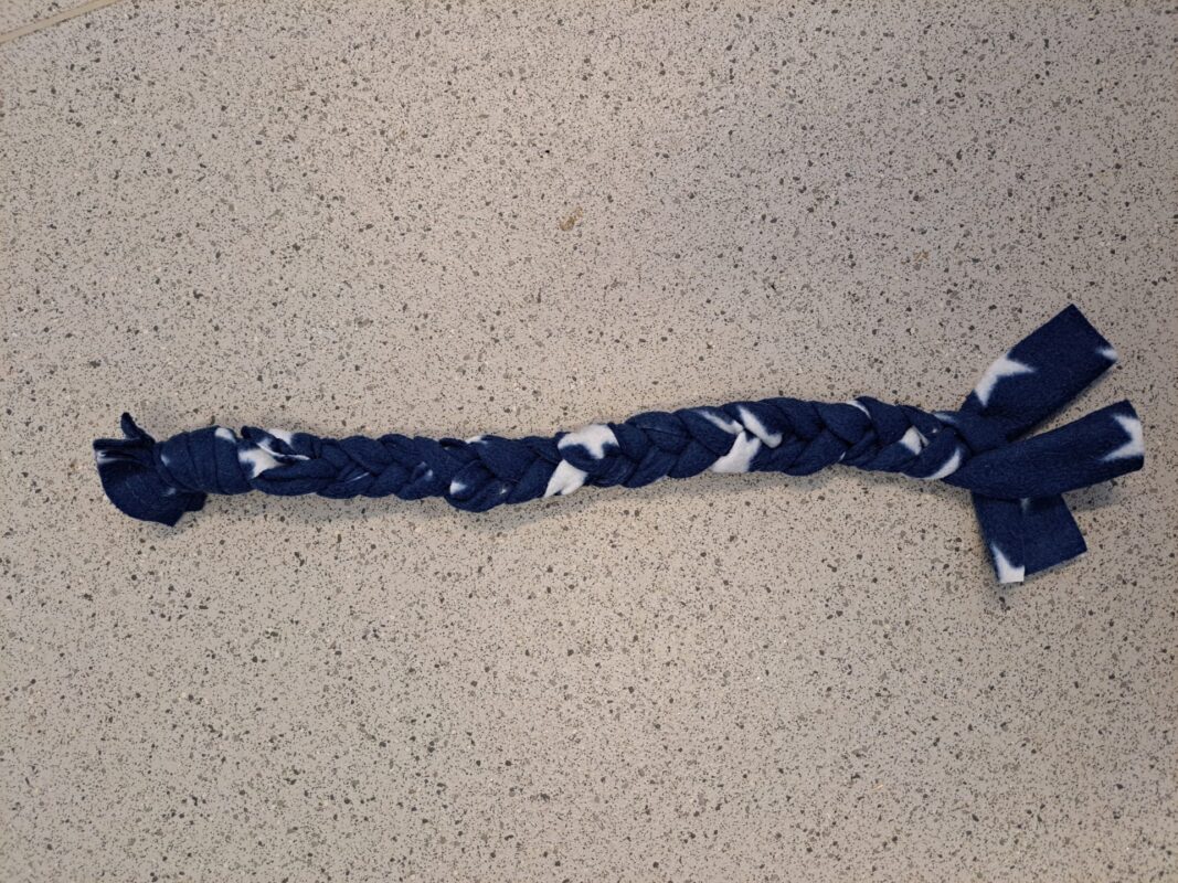 A braided dog toy made of blue and white fleece fabric rests on a speckled countertop. Similar to a rat hammock, the toy features knotted ends and loose fabric strips at the bottom.