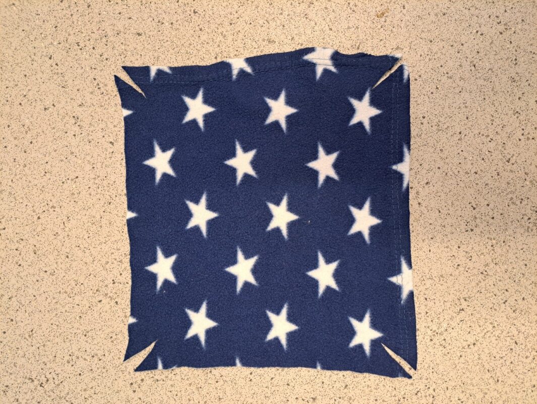 A square rat hammock of dark blue fabric with white stars rests on a speckled surface. Each corner of the hammock features a short diagonal cut.