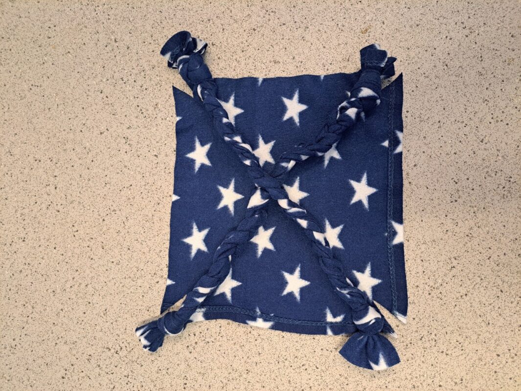 A square rat hammock made of navy blue fleece with white stars, featuring four braided corners tied together diagonally, placed on a speckled beige countertop.