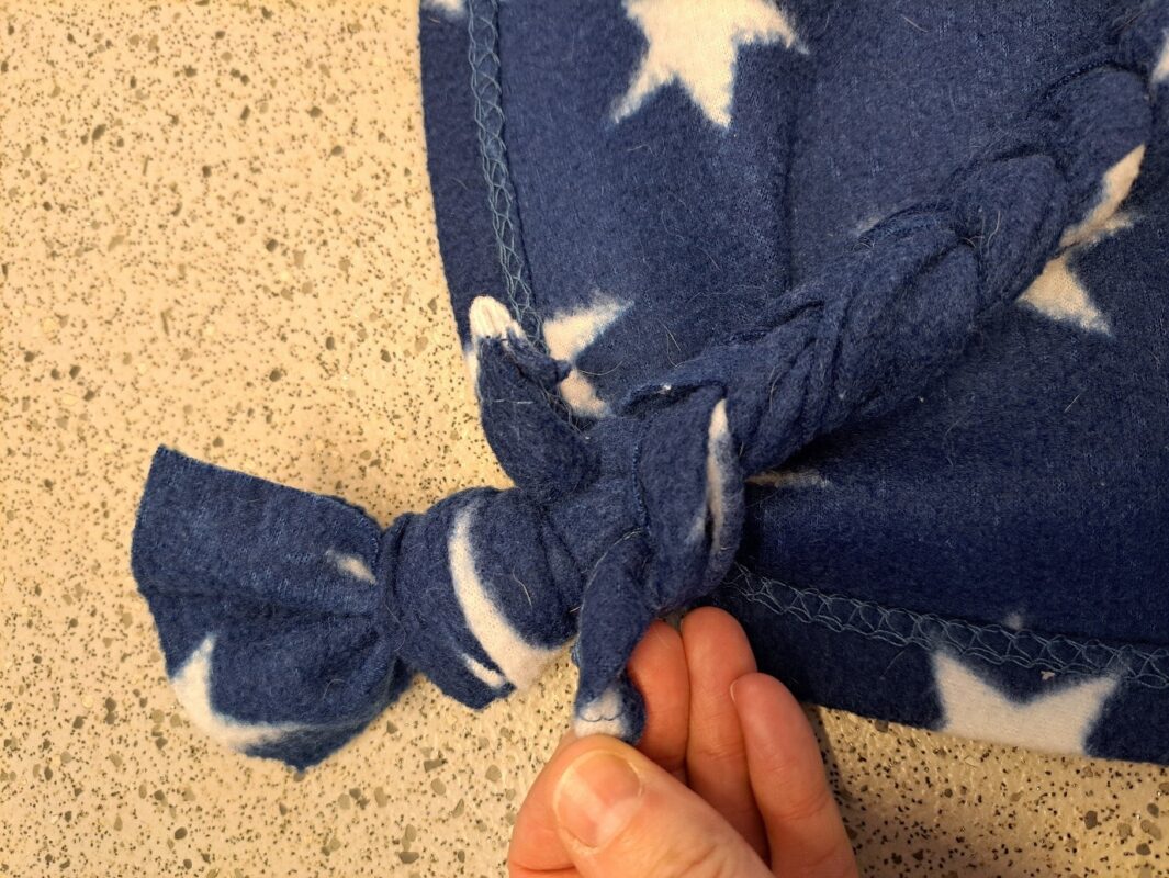 A hand holding the knotted edge of a blue fleece rat hammock with white stars, displaying its braided and tied section on a speckled countertop.