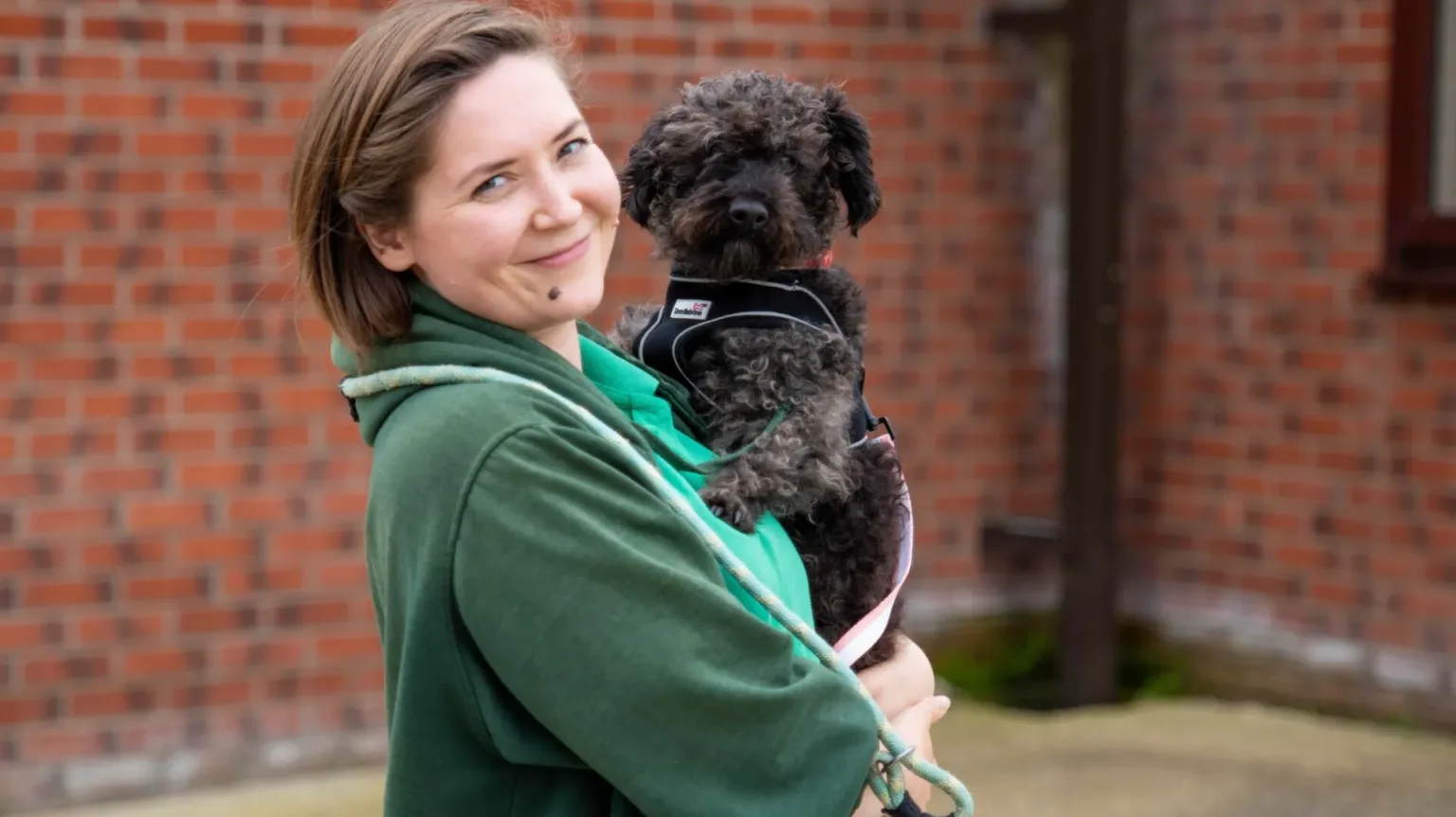 A black curly haired Terrier is held in the arms of a Woodgreen staff member. They are both looking at the camera.