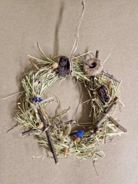 A small, rustic wreath for rabbits and rodents, made of dried grass, twigs, and wildflowers, with a loop of string for hanging, displayed on a plain brown background.