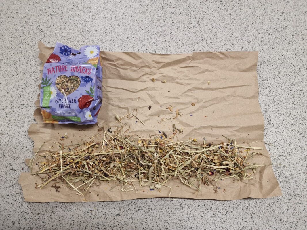 A packet of Nature Snacks wildflower forage is open on a speckled countertop, with an assortment of dried hay, flowers, and herbs spread out on a sheet of brown paper beside it.