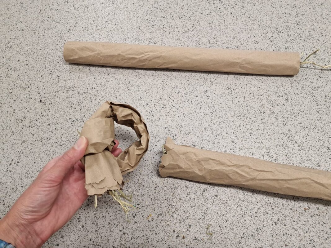 A hand holds a partially rolled ring of brown paper filled with straw, with two long, rolled paper tubes filled with straw lying on a speckled countertop.