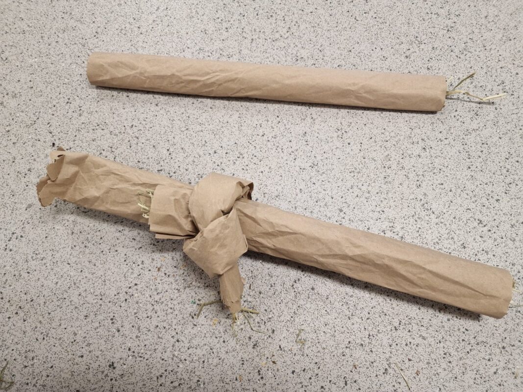 Two rolled up tubes wrapped of brown paper sit on a speckled countertop. One is plain, while the other has a knot tied in the middle of the paper, with some shredded material sticking out of the ends.