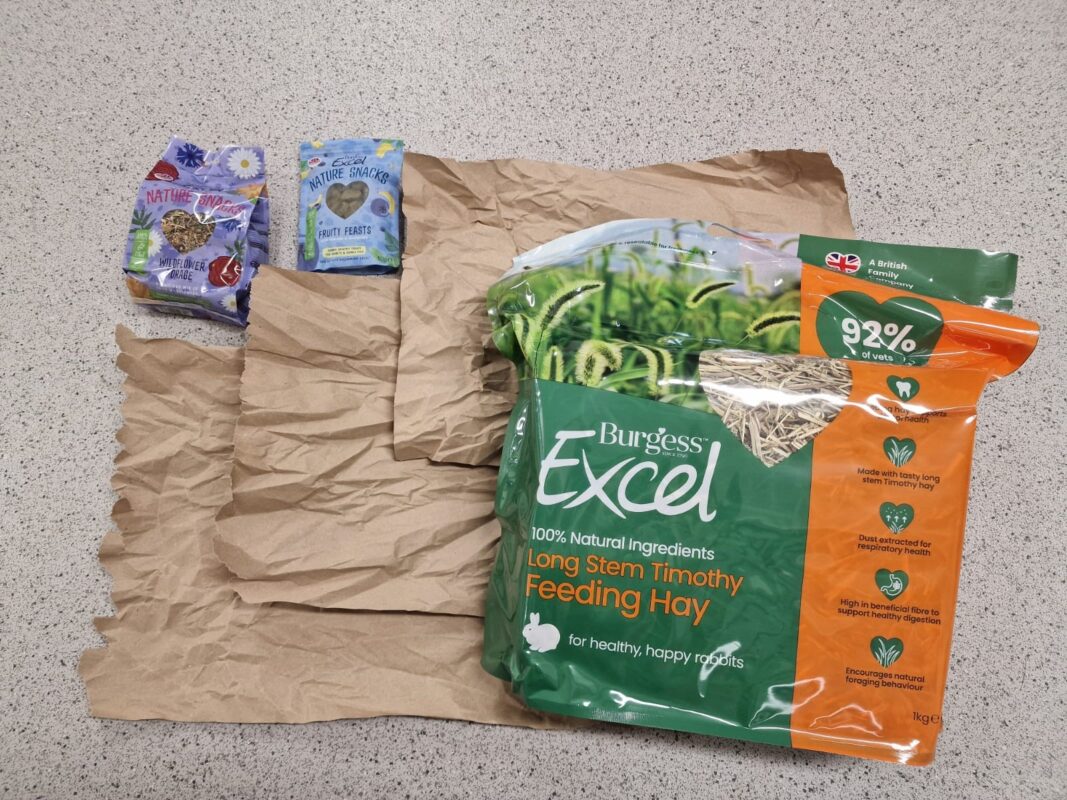 A bag of Burgess Excel Long Stem Timothy Feeding Hay is on a counter next to two small packets of rodent food and two pieces of brown paper.