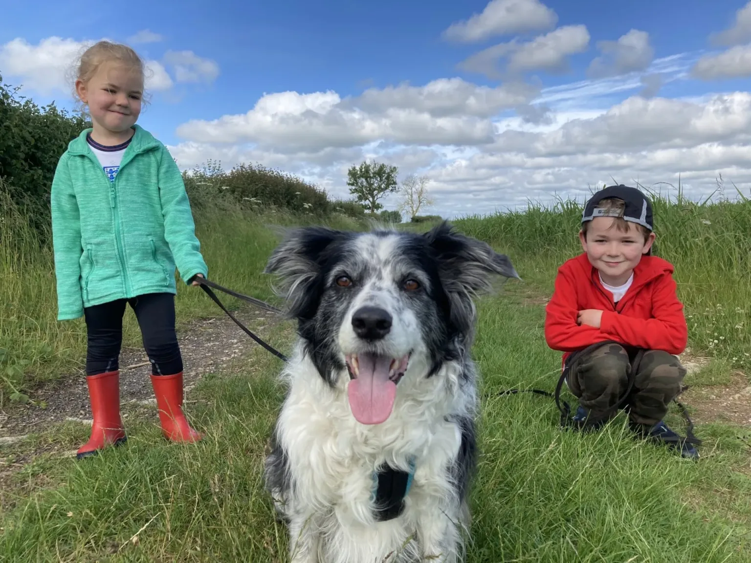 Two smiling children, one in a turquoise jacket and red boots, and one in a red hoodie and cap, pose outdoors on a grassy path with a happy black and white dog in the center, under a partly cloudy sky.