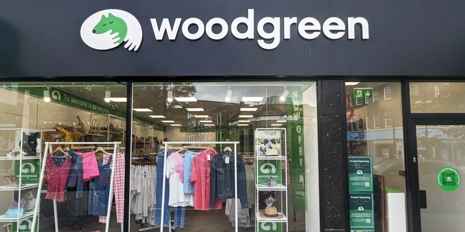 Front view of a Woodgreen charity shop, featuring large logo signage above the entrance and display racks with colorful clothing visible through the windows. Green grand opening signs are on the door.
