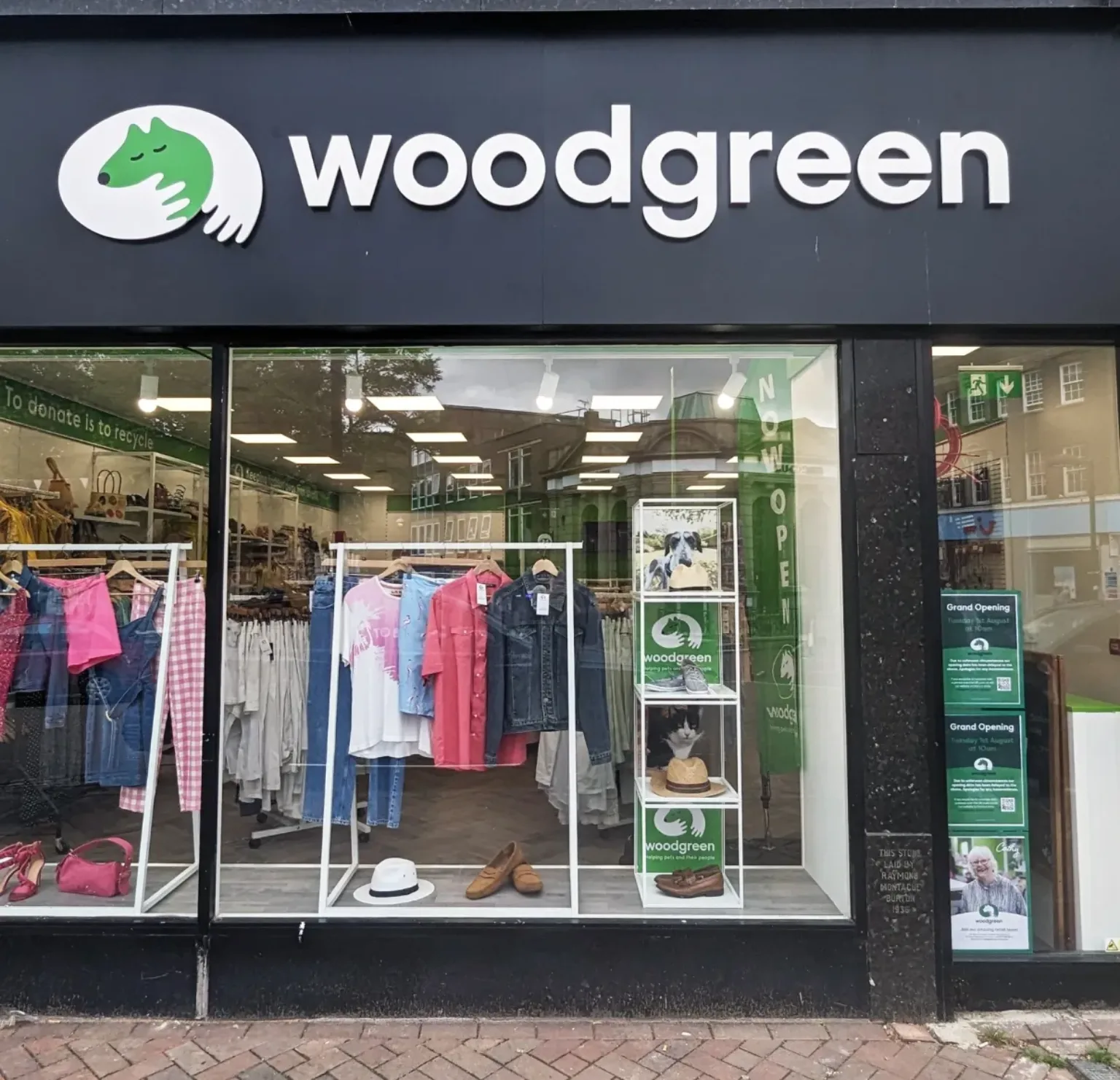 The storefront of a Woodgreen charity shop displays clothing, shoes, hats, and accessories in the window. The Woodgreen logo and name are above the entrance, and posters about the grand opening are on the glass.