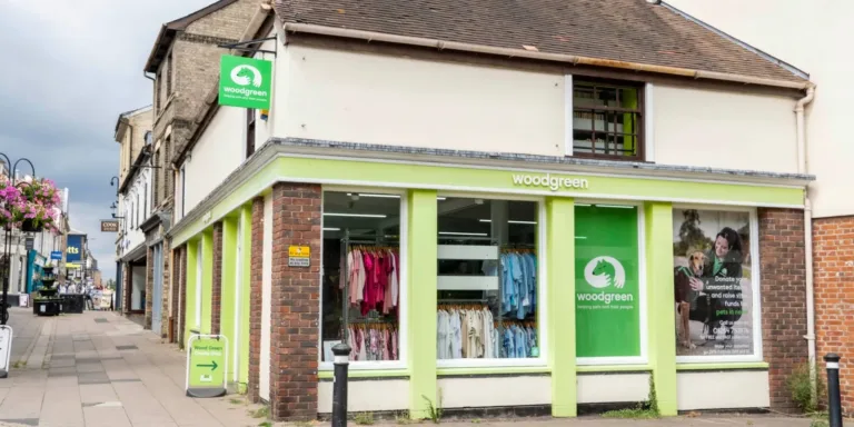 A corner shop with woodgreen signage and a green logo, large display windows showing clothes inside, and a poster with a woman and a dog. The building has brick and light green trim. The street is quiet.