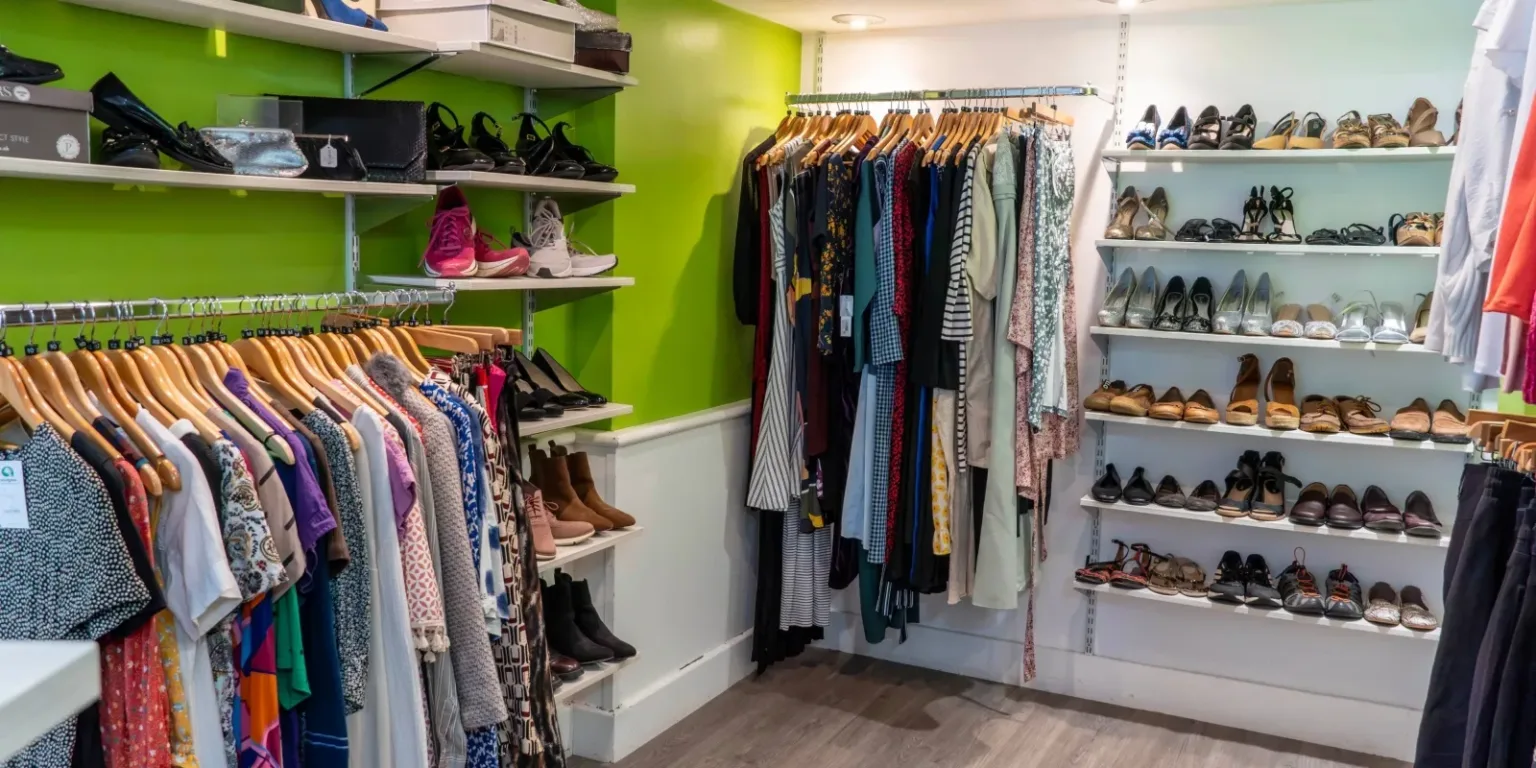 A brightly lit clothing and shoe boutique features colorful dresses and shirts hanging on racks, with shelves displaying various shoes, sandals, and boots against green and white walls.