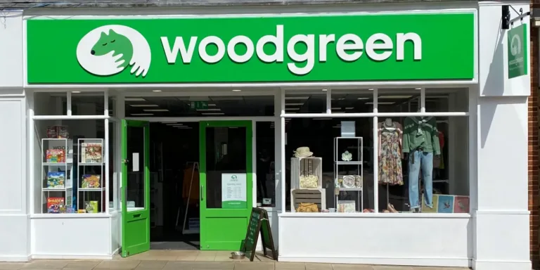 A storefront with a green and white sign reading woodgreen and a logo of a dog and hands. The shop has glass windows displaying clothing, books, and various items. The door is open, inviting customers inside.
