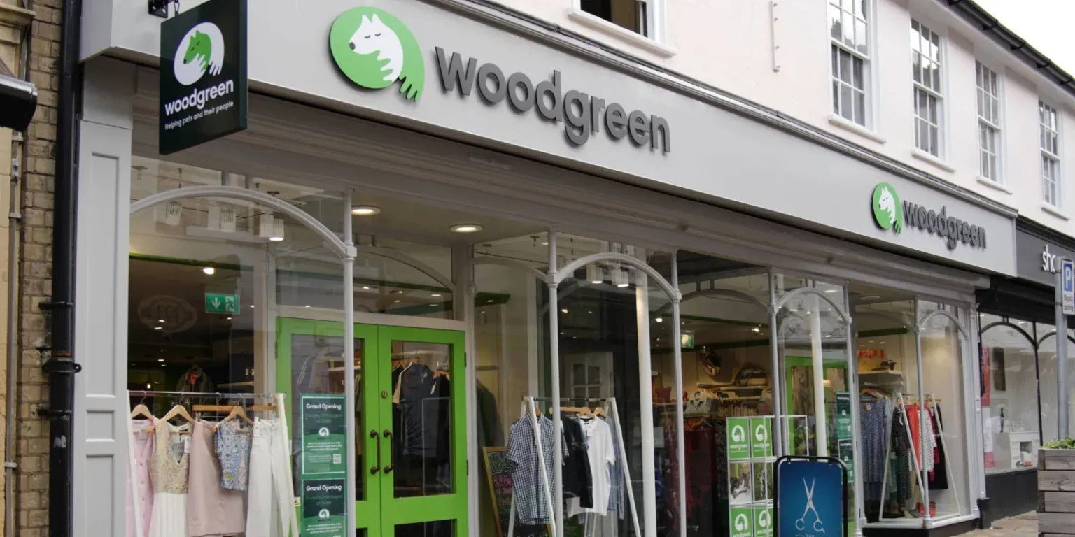 The storefront of a Woodgreen charity shop, with large windows displaying clothing and a green door. The Woodgreen logo and name are visible above the entrance and on a hanging sign.