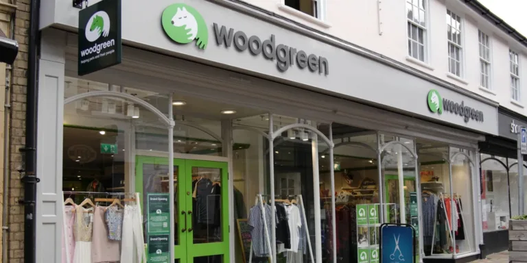 The storefront of a Woodgreen charity shop, with large windows displaying clothing and a green door. The Woodgreen logo and name are visible above the entrance and on a hanging sign.